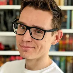 Uśmiechnięty mężczyzna w okularach na tle regałów z książkami. Może analizuje przyczyny fascynacji pisarzy danymi statystycznymi dotyczącymi ich twórczości.