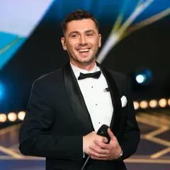 Kamil Bednarek w czarnym smokingu z muszką, trzyma mikrofon i uśmiecha się.