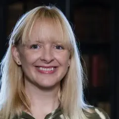 Uśmiechnięta blondynka z grzywką, w tle biblioteka. Jej spojrzenie sugeruje, jakie są relacje między autorem a jego literackim alter ego – pełne refleksji.