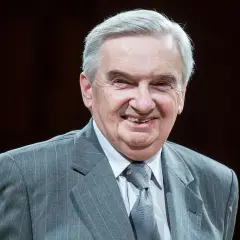 Uśmiechnięty starszy pan w szarym garniturze. Trudno zgadnąć, ile lat ma sznuk, ale wygląda na doświadczonego i życzliwego.