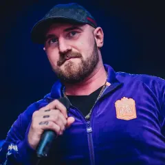 Kizo w fioletowej bluzie z kapturem i czapce, trzymający mikrofon. Fani zastanawiają się, ile wzrostu ma Kizo.