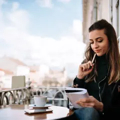 Młoda kobieta z notatnikiem i kawą zastanawia się, czy warto publikować najpierw na platformach typu Wattpad czy Royal Road.