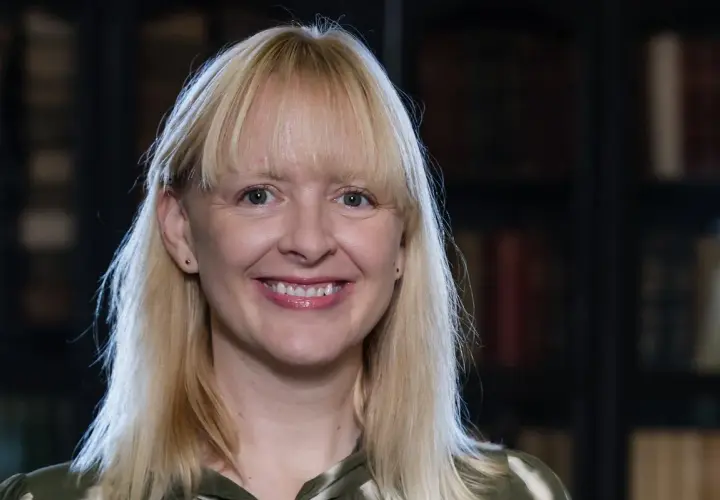 Uśmiechnięta blondynka z grzywką, w tle biblioteka. Jej spojrzenie sugeruje, jakie są relacje między autorem a jego literackim alter ego – pełne refleksji.