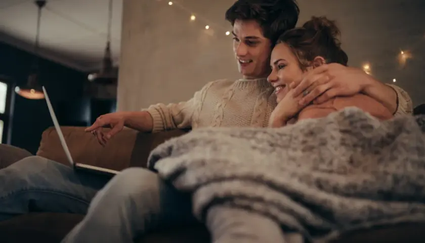 Para młodych ogląda coś na laptopie, przytuleni pod kocem. Ich bliskość i uśmiechy sugerują, że historia o "fake dating" tak bardzo angażuje emocjonalnie czytelników, bo pozwala przeżywać rodzące się uczucia.