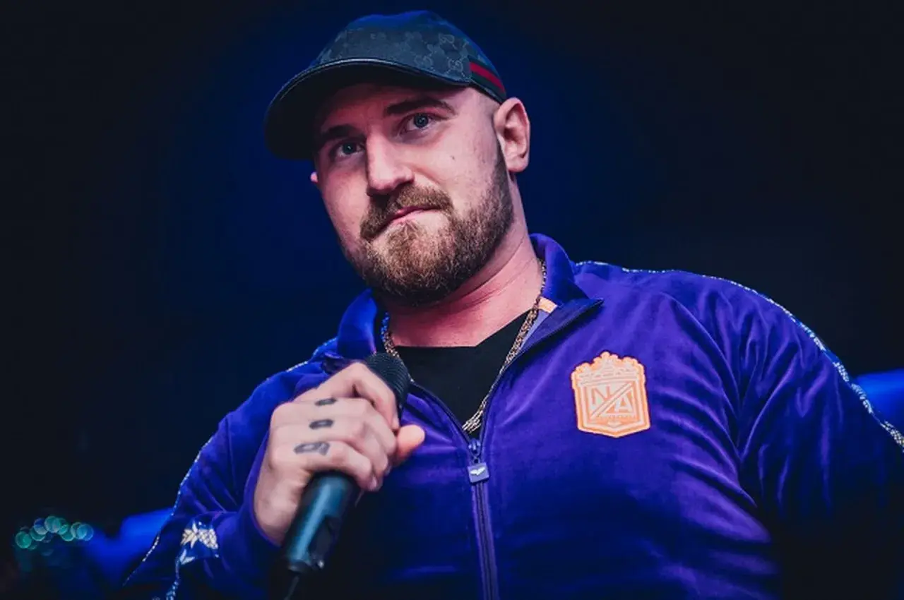 Kizo w fioletowej bluzie z kapturem i czapce, trzymający mikrofon. Fani zastanawiają się, ile wzrostu ma Kizo.