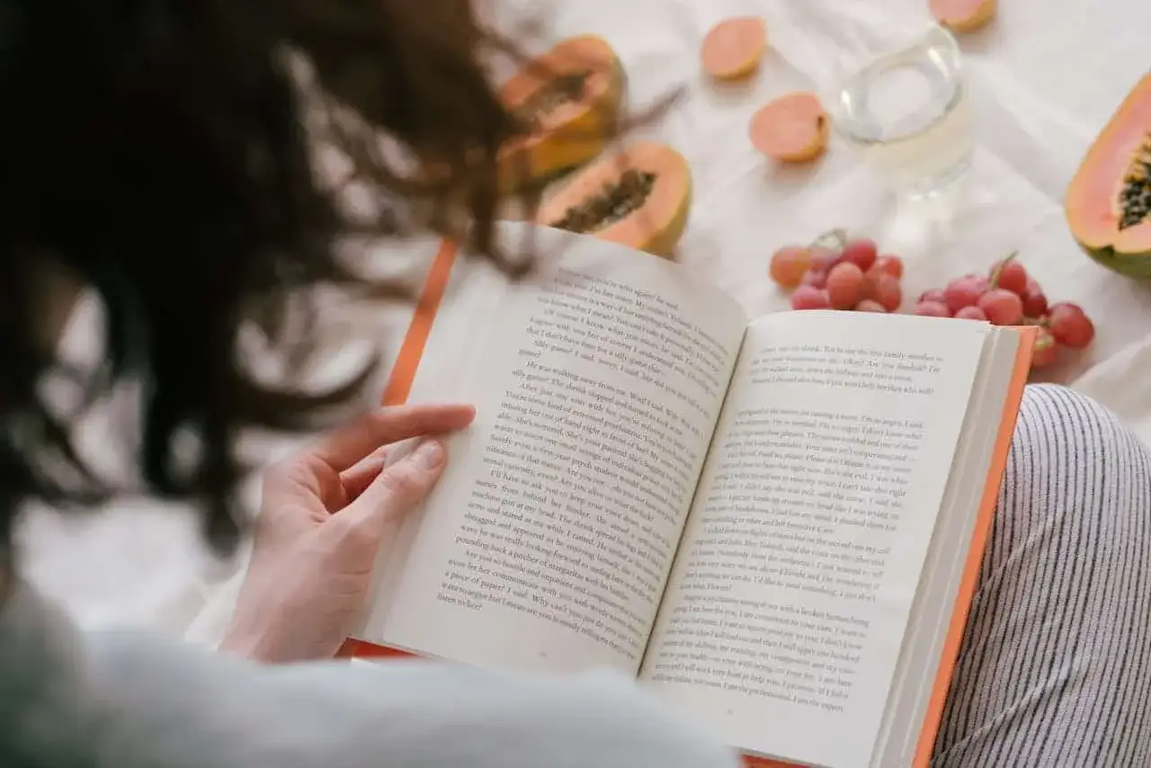 Osoba czyta książkę, zastanawiając się, czy audiobooki zmieniają sposób, w jaki odbieramy świat przedstawiony. Na stole owoce i szklanka.