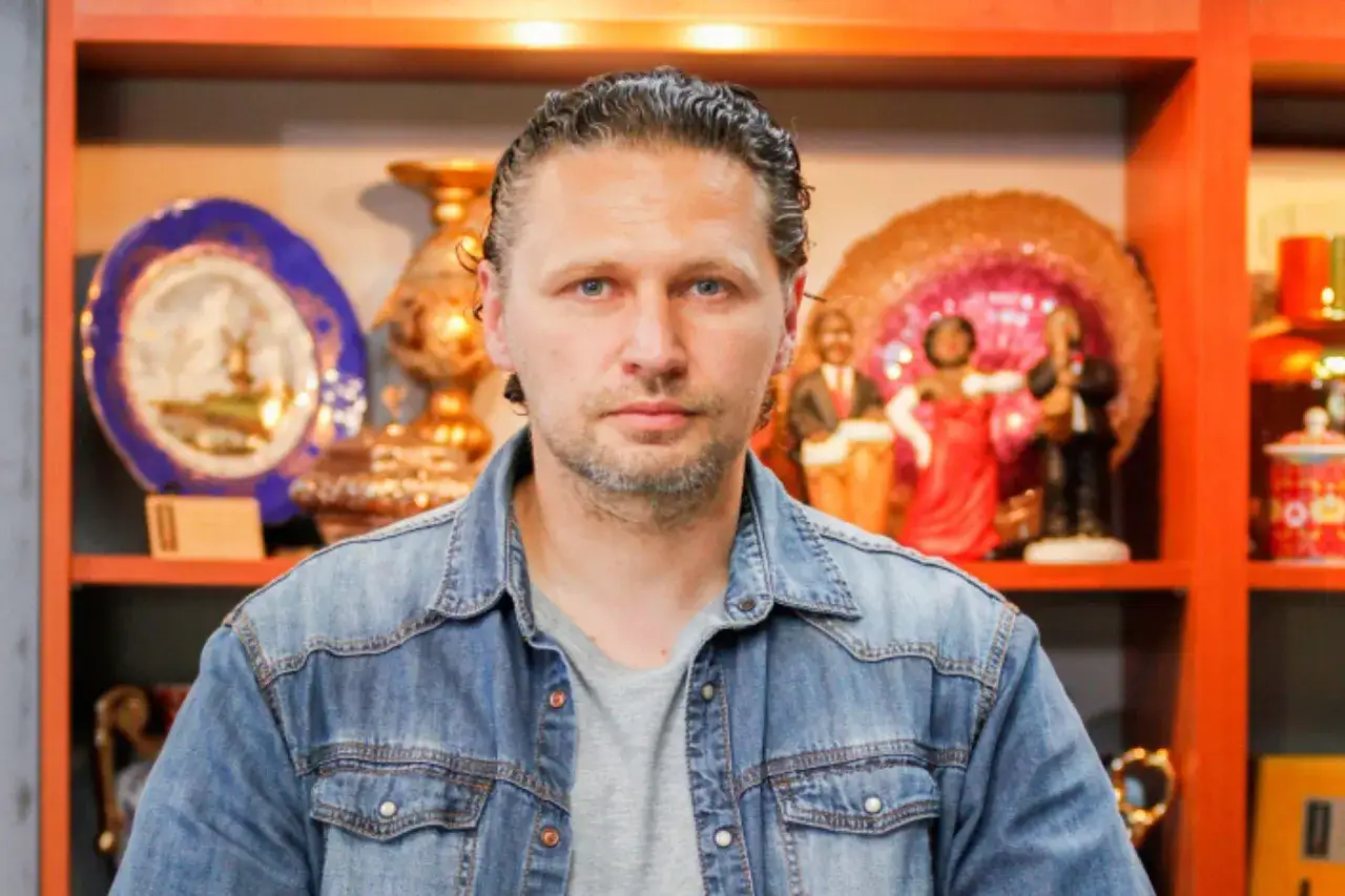 Mariusz z lombardu, w dżinsowej koszuli, stoi na tle półek z bibelotami. Ile lat ma Mariusz z lombardu?